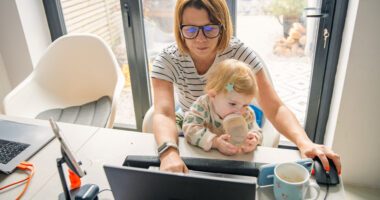 Woman sits at kitchen table, working on laptop, while toddler sits in her lap