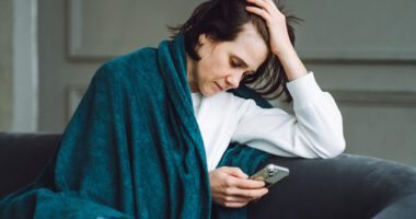 woman wears a blanket over her shoulders, hand on her forehead, looking on her phone