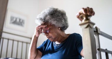 older woman sits on bed, frustrated, hand resting on bed post for support