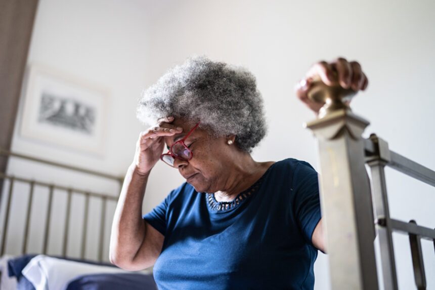 older woman sits on bed, frustrated, hand resting on bed post for support