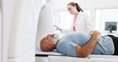 In the image, a man is lying inside a CT scan machine while a healthcare professional stands beside him, operating the equipment and monitoring the procedure.