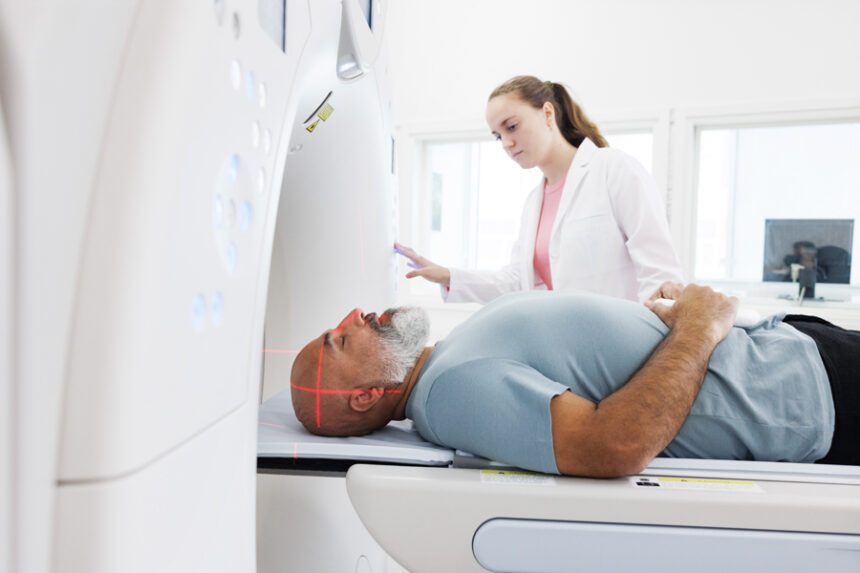 In the image, a man is lying inside a CT scan machine while a healthcare professional stands beside him, operating the equipment and monitoring the procedure.