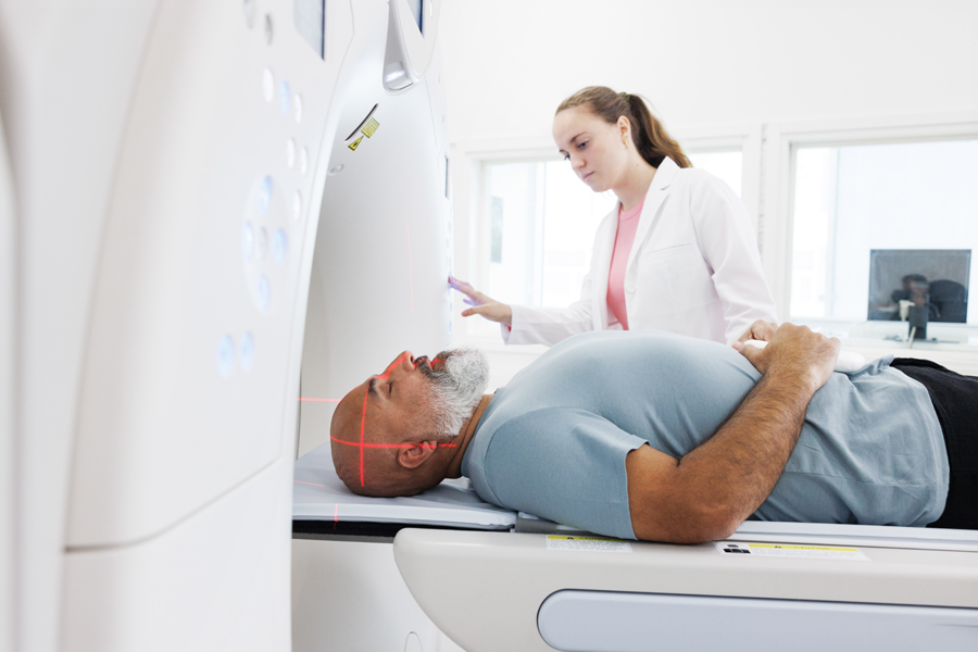 In the image, a man is lying inside a CT scan machine while a healthcare professional stands beside him, operating the equipment and monitoring the procedure.
