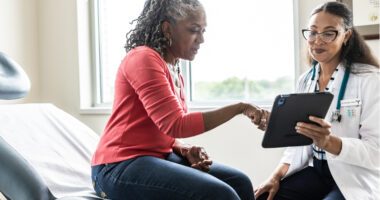A patient is sitting on an examination table in a gynecological office, while a gynecologist, seated beside her, holds a digital tablet.