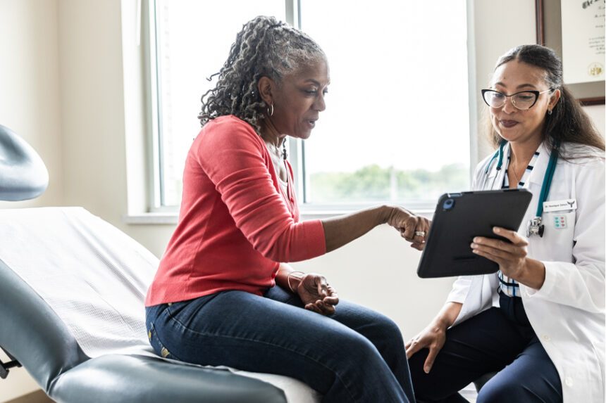 A patient is sitting on an examination table in a gynecological office, while a gynecologist, seated beside her, holds a digital tablet.