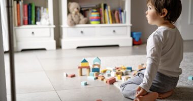 A young child is sitting on the floor, staring off into the distance while playing alone with colorful building blocks in a neatly organized children's room.
