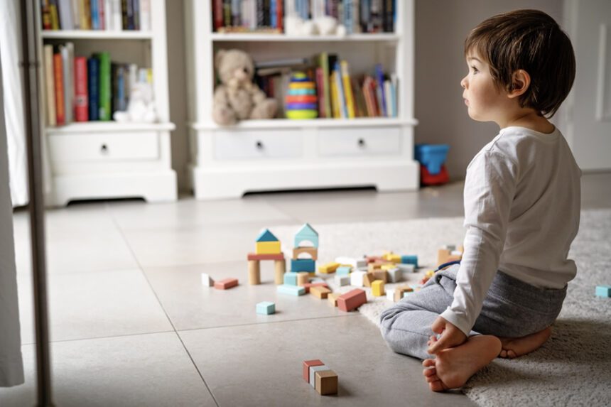 A young child is sitting on the floor, staring off into the distance while playing alone with colorful building blocks in a neatly organized children's room.