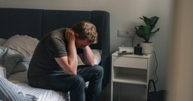 A man sitting on the edge of a bed, leaning forward with his hands behind his neck, looking distressed or worried.