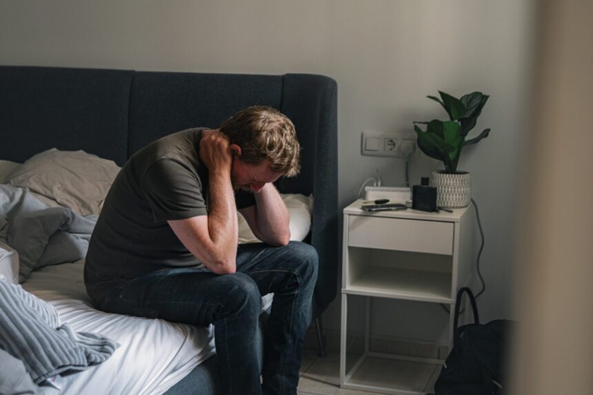 A man sitting on the edge of a bed, leaning forward with his hands behind his neck, looking distressed or worried.