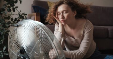 young woman sits in front of a metal cooling fan, eyes closed.
