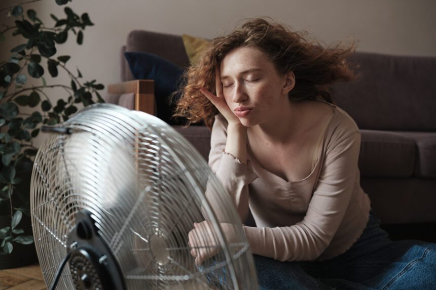 young woman sits in front of a metal cooling fan, eyes closed.