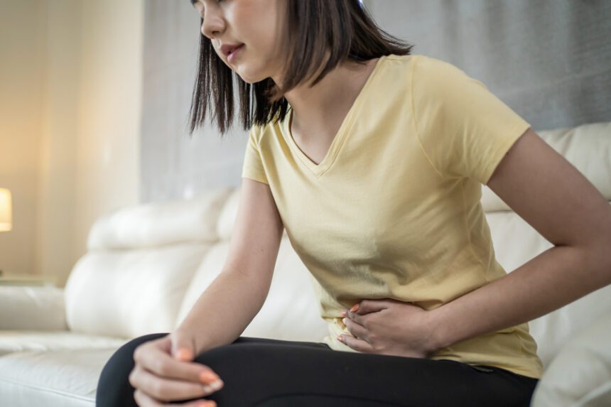 A woman sitting on a couch holding her lower abdomen, appearing to be in pain or discomfort.
