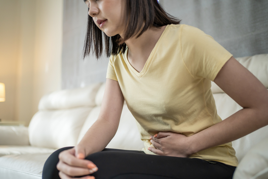 A woman sitting on a couch holding her lower abdomen, appearing to be in pain or discomfort.