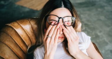 A woman wearing glasses is rubbing her eyes with both hands while sitting in a chair, looking tired or uncomfortable.