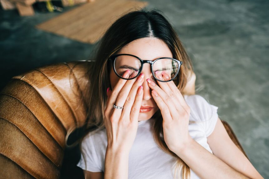A woman wearing glasses is rubbing her eyes with both hands while sitting in a chair, looking tired or uncomfortable.