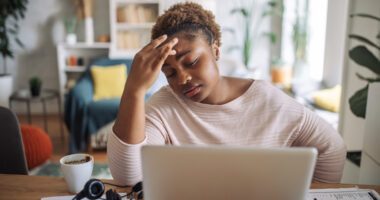 A woman looks visibly fatigued while working on her laptop, with one hand on her forehead and an expression of exhaustion in a home setting.