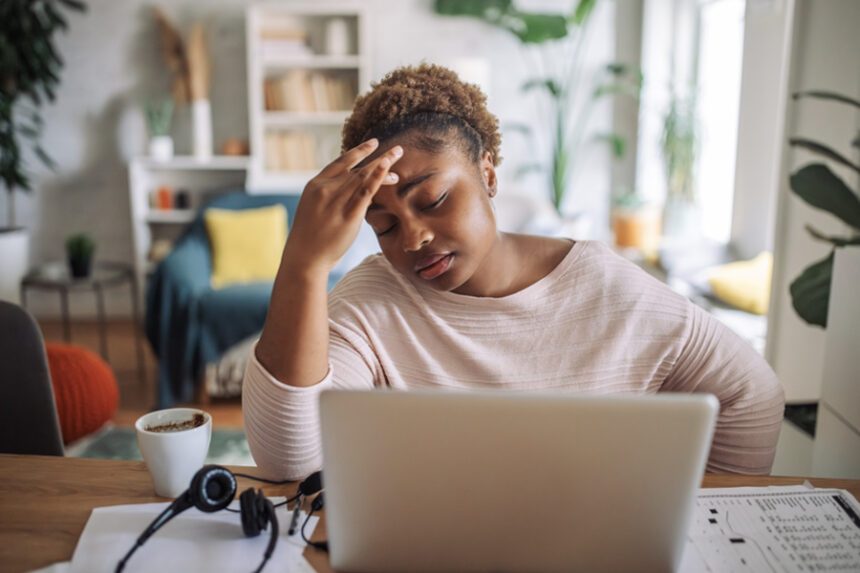 A woman looks visibly fatigued while working on her laptop, with one hand on her forehead and an expression of exhaustion in a home setting.