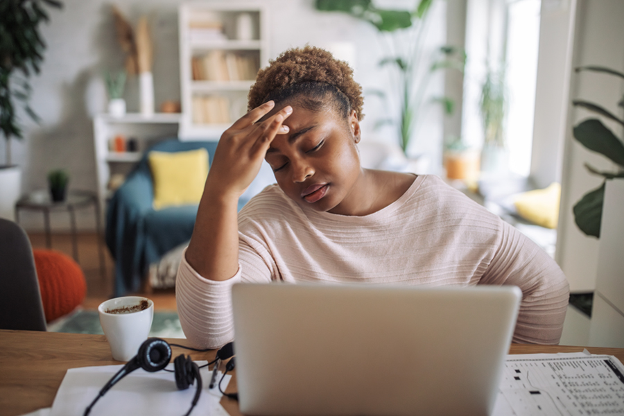 A woman looks visibly fatigued while working on her laptop, with one hand on her forehead and an expression of exhaustion in a home setting.