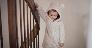 A little girl, dressed in a teddy bear-style onesie, calmly walks down the stairs while holding onto the handrail.