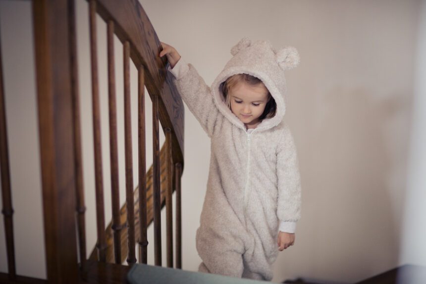 A little girl, dressed in a teddy bear-style onesie, calmly walks down the stairs while holding onto the handrail.