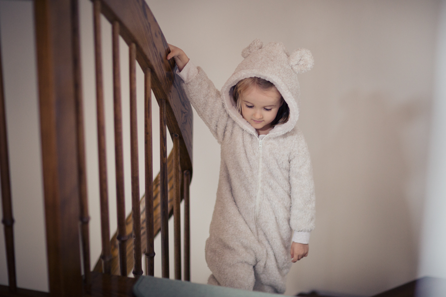 A little girl, dressed in a teddy bear-style onesie, calmly walks down the stairs while holding onto the handrail.