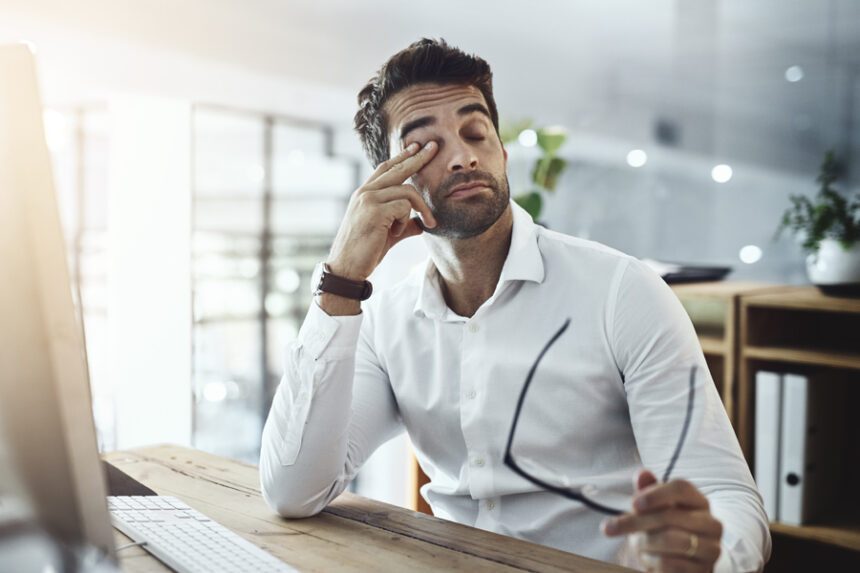 A tired-looking man rubs one eye while holding his glasses, sitting at a desk in an office setting.