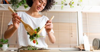 A smiling young woman prepares a fresh salad in a bright kitchen, enjoying a moment of healthy well-being.