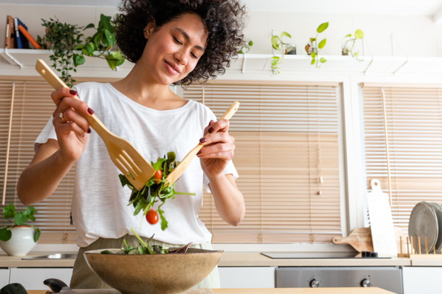 A smiling young woman prepares a fresh salad in a bright kitchen, enjoying a moment of healthy well-being.