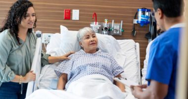An elderly patient lying in a hospital bed speaks with a healthcare professional while a family member stands by her side.