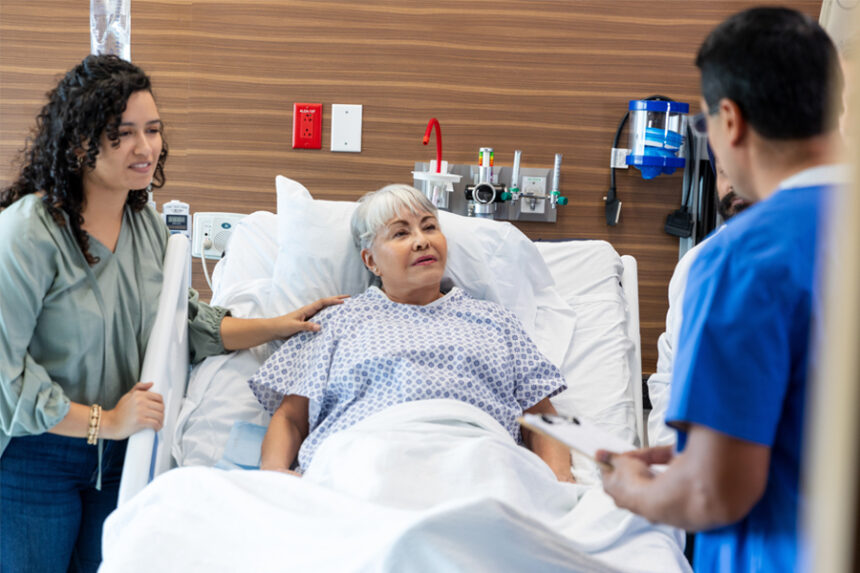 An elderly patient lying in a hospital bed speaks with a healthcare professional while a family member stands by her side.