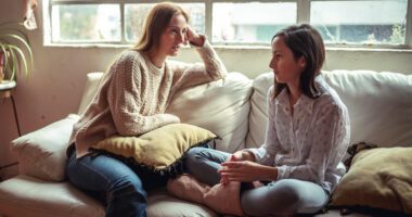 A woman is having a close and attentive conversation with a teenager, sitting on a couch in a cozy setting by a window.