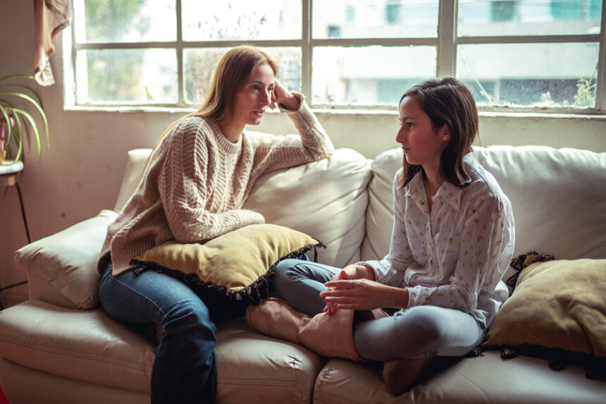 A woman is having a close and attentive conversation with a teenager, sitting on a couch in a cozy setting by a window.