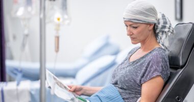 A patient wearing a headscarf sits in a treatment chair receiving chemotherapy while reading a magazine.