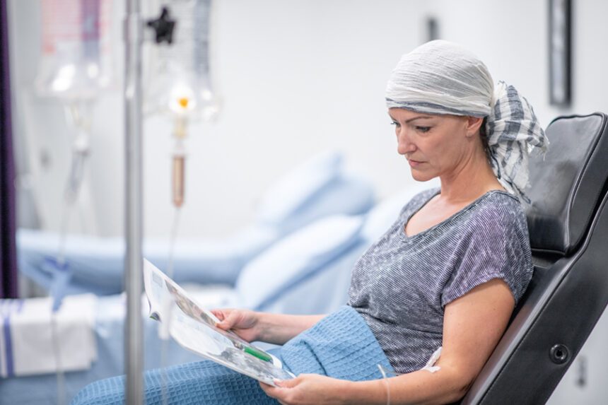 A patient wearing a headscarf sits in a treatment chair receiving chemotherapy while reading a magazine.