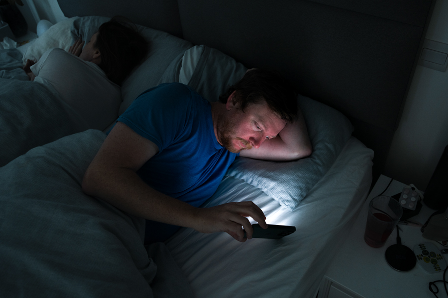 A man lying in bed, unable to sleep, looks at his phone while his partner sleeps beside him.