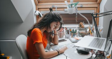 A young woman sits at her desk with her head resting on her hand, looking tired or stressed while studying or working on her laptop.