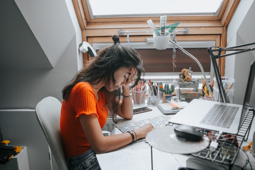 A young woman sits at her desk with her head resting on her hand, looking tired or stressed while studying or working on her laptop.
