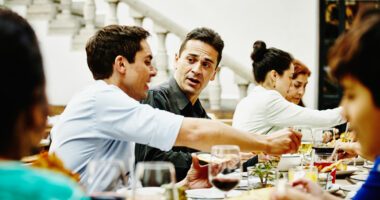 A group of adults sit around a dinner table, engaged in a tense conversation that suggests interpersonal conflict during a family gathering.