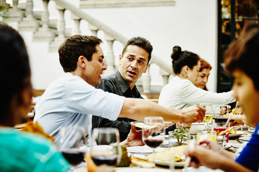 A group of adults sit around a dinner table, engaged in a tense conversation that suggests interpersonal conflict during a family gathering.