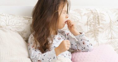 A young girl sits on a couch, coughing into her hand.