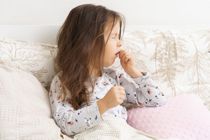 A young girl sits on a couch, coughing into her hand.