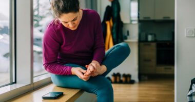 woman sits on window seat in home, examining the bottom of her foot