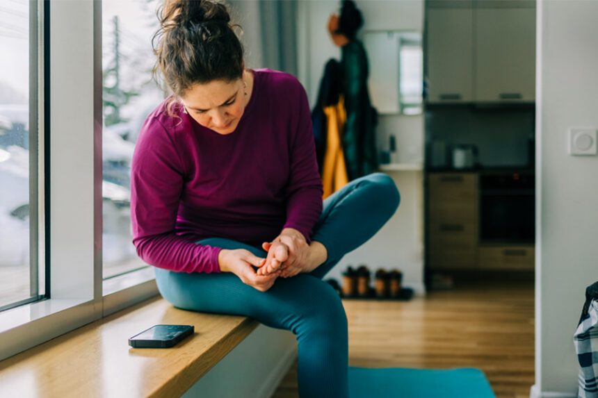 woman sits on window seat in home, examining the bottom of her foot