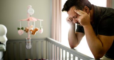 Father leans over a crib, massaging temples in look of stress