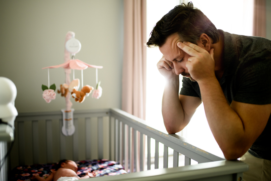 Father leans over a crib, massaging temples in look of stress