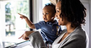 Mom holds baby while they look out of a window, pointing at objects they see