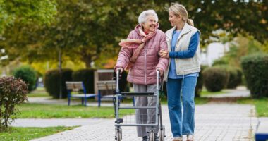 Elderly woman walks with walker, a caretakers walks by her side, smiling