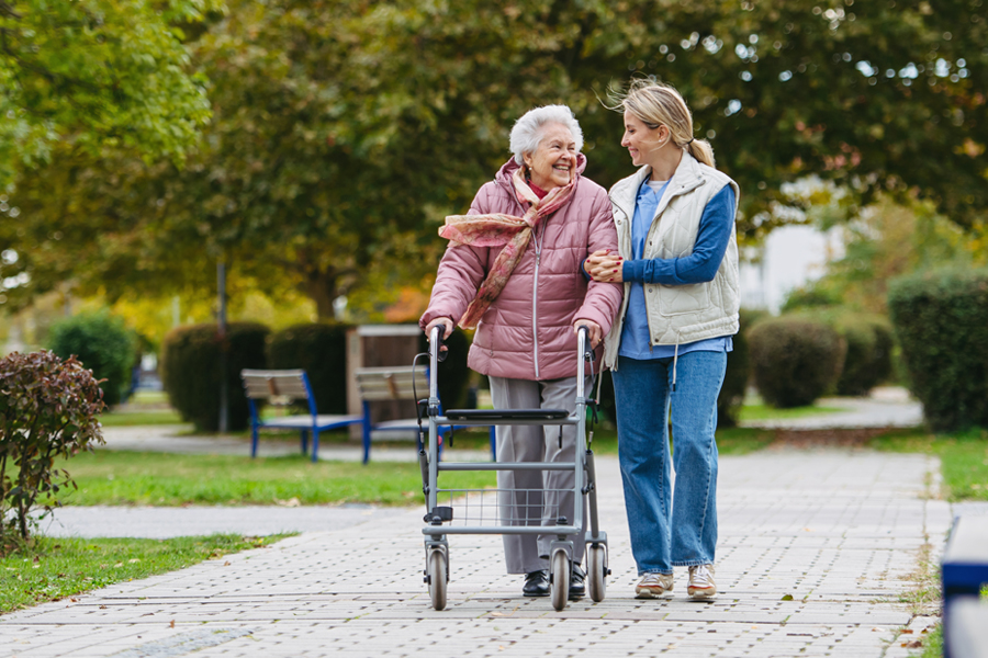 Elderly woman walks with walker, a caretakers walks by her side, smiling