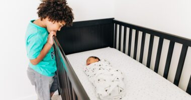 older sibling looks at baby sibling swaddled in a crib