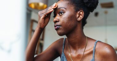 An adult examines acne on her face in the mirror
